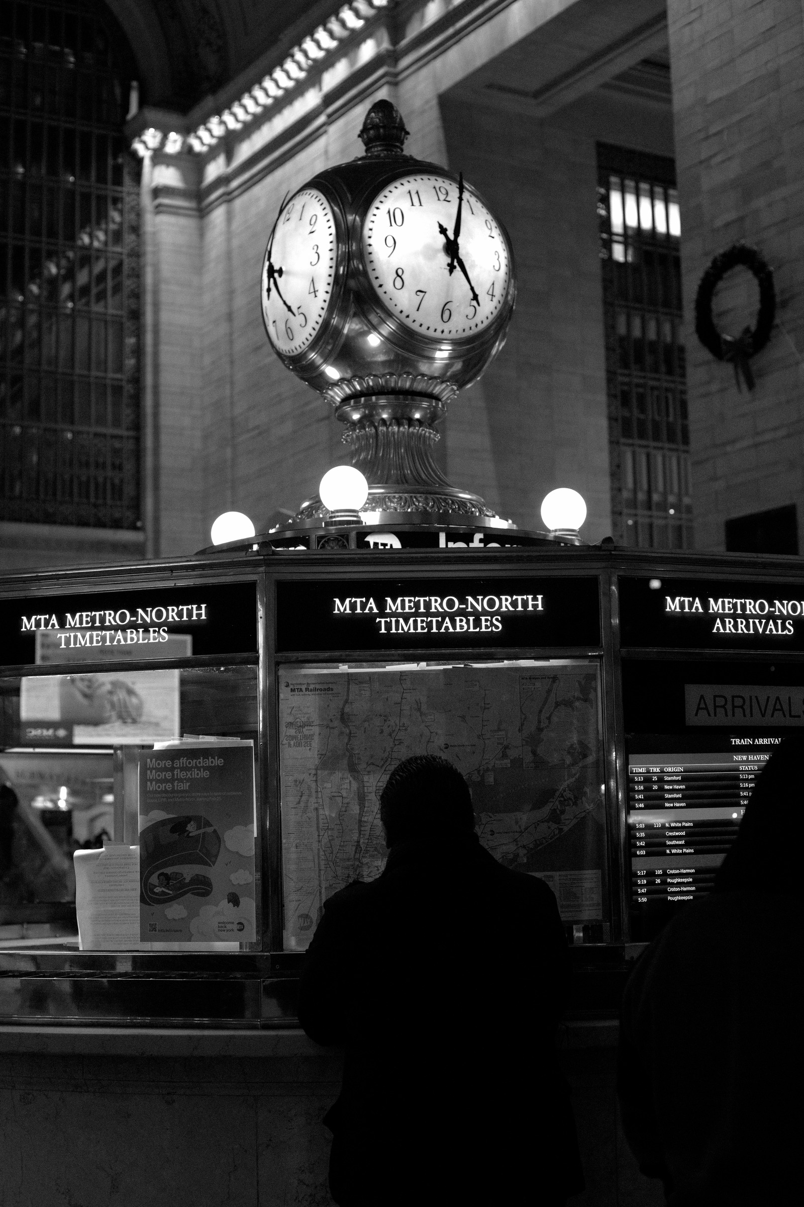 A silhouetted passenger beneath the clock at a railway information booth.